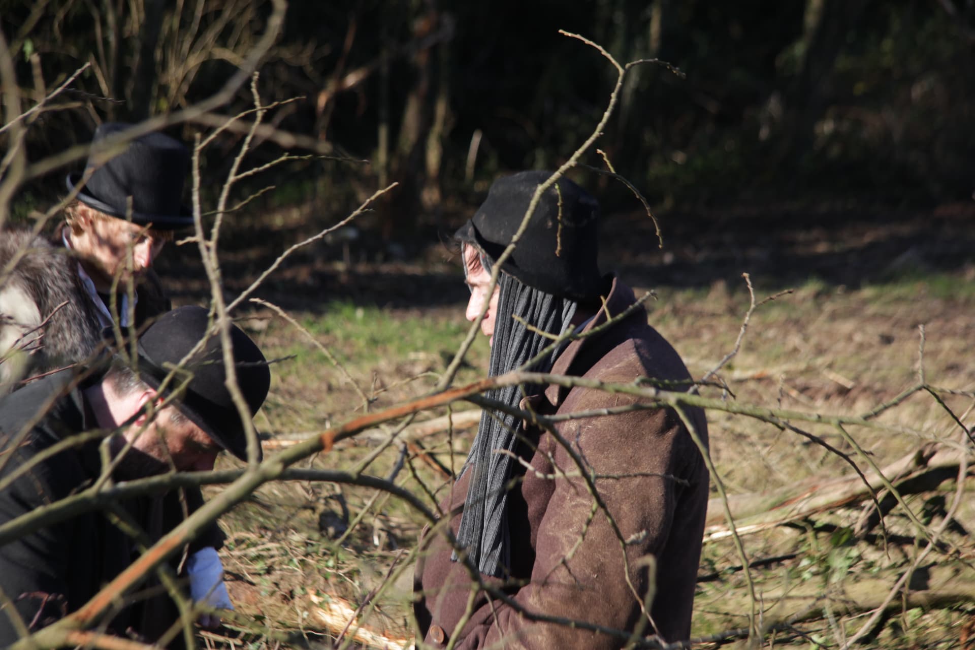 Two actors in period hats and coats waiting in the hedgerow